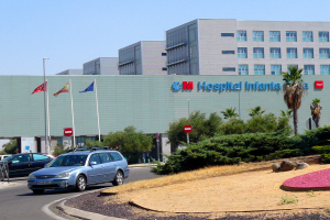 Krankenhaus mit geparkten Autos, umgeben von städtischen Elementen einschließlich Gebäuden, Straßeninfrastruktur, Fahrzeugen, Grünflächen und einem klaren Himmel.