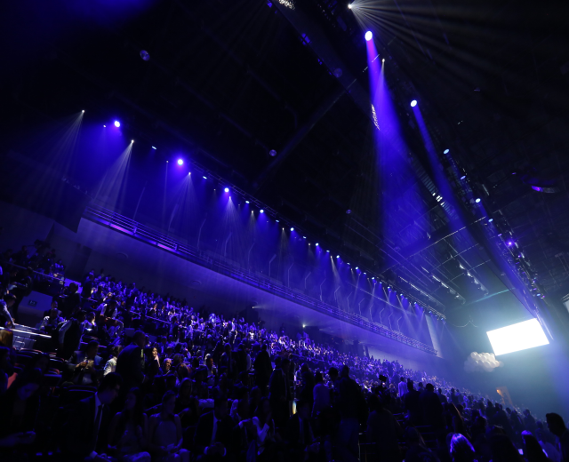 Eine große Menschenmenge, die in einem Konzertsaal auf einer Bühne sitzt.