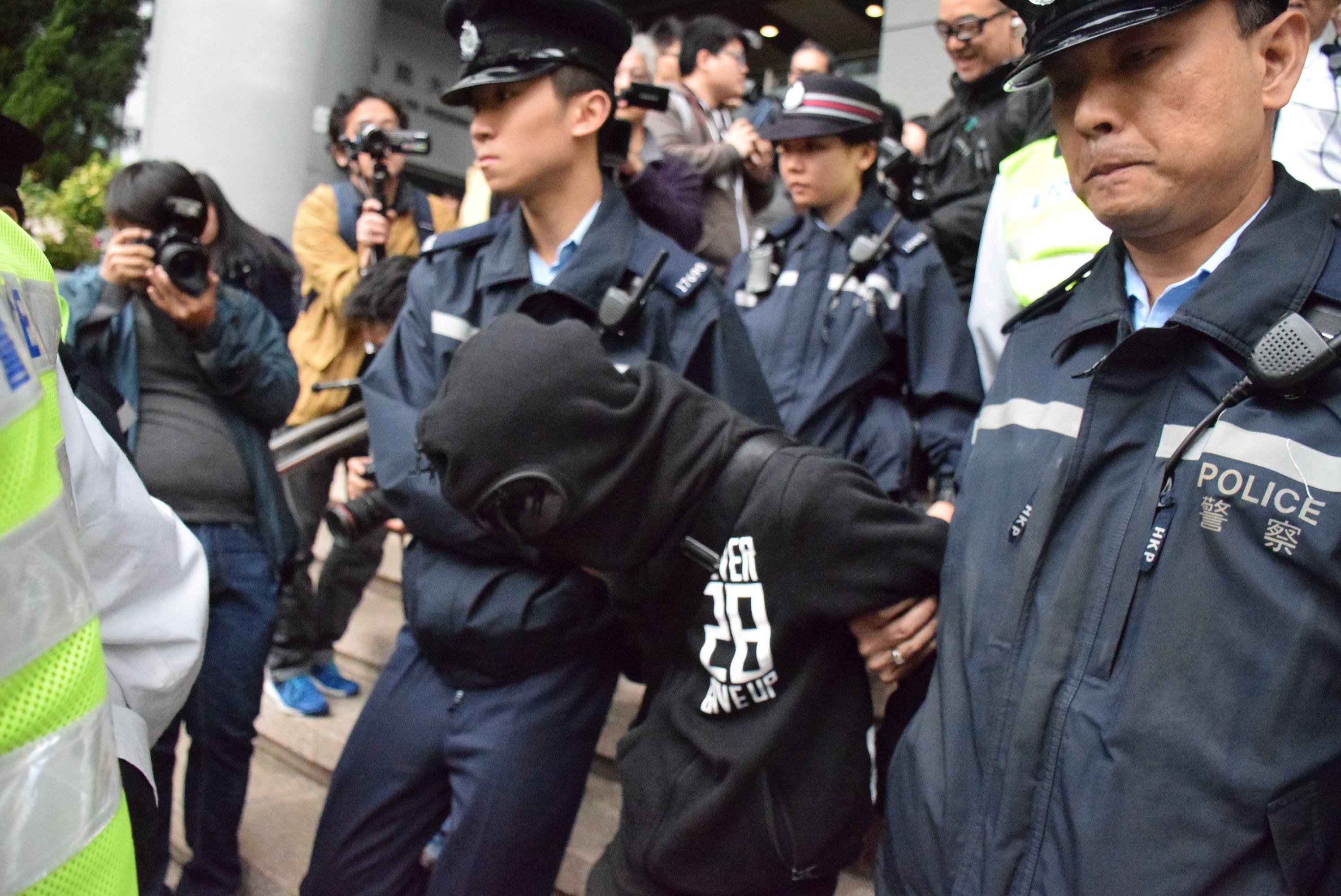 Eine Gruppe von Polizisten steht vor einer Menschenmenge, einige tragen Mützen und halten Kameras, im Hintergrund ein Gebäude mit Säulen und Deckenleuchten und links Bäume.