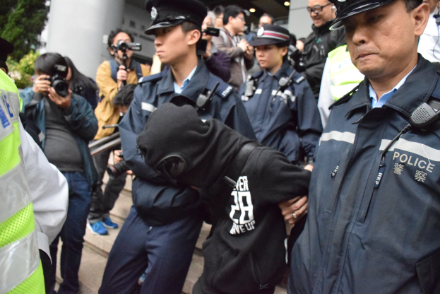 Eine Gruppe von Polizisten steht vor einer Menschenmenge, einige tragen Mützen und halten Kameras, im Hintergrund ein Gebäude mit Säulen und Deckenleuchten und links Bäume.