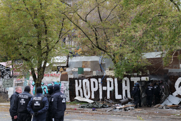 Gruppe von Polizisten in schwarzen Uniformen und Helmen vor einem mit Graffiti bedeckten, baufälligen Gebäude mit verstreuten Trümmern und Bäumen im Hintergrund.