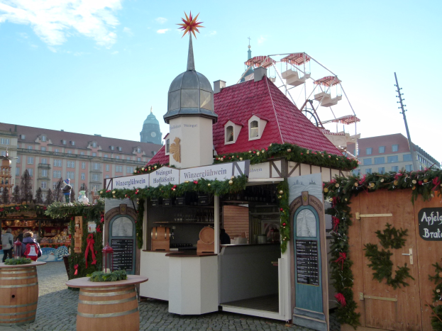 Ein kleines Gebäude mit einem roten Dach und einem Riesenrad davor, umgeben von Menschen und Festschmuck, vor einer Kulisse aus Gebäuden, Bäumen und einem bewölkten Himmel.
