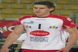 Ein Mann in einem roten und weißen Handballtrikot steht auf einem Feld, umgeben von Zuschauern, mit einem roten Banner im Hintergrund, auf dem "Krasnoyarsk vs Bydgoszcz" steht, und sieht entschlossen aus.