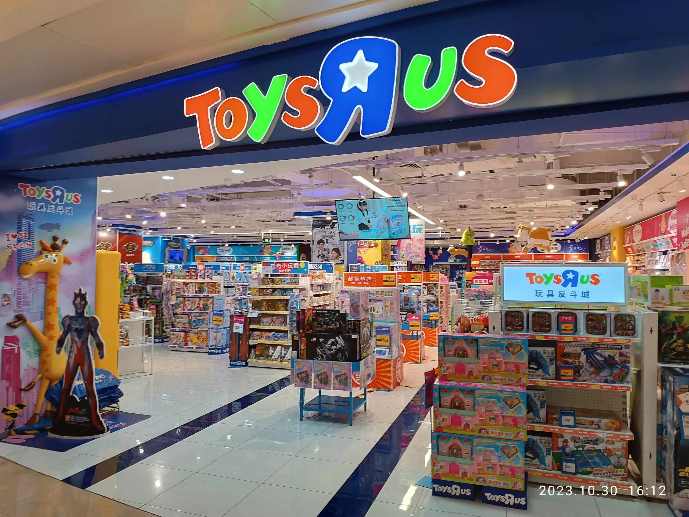 Ein Toys R Us-Laden in einem Einkaufszentrum, der Regale mit Spielzeug und anderen Artikeln zeigt, ein sichtbares Ladenschild, Deckenbeleuchtung und ein Wasserzeichen in der rechten unteren Ecke.
