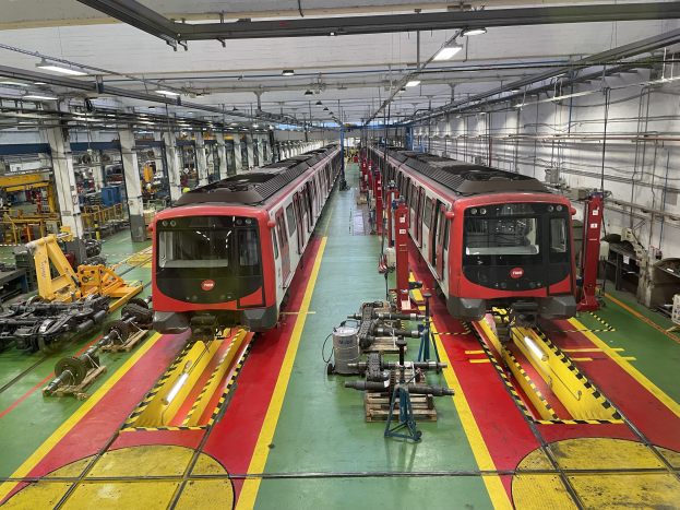 Zwei Züge auf Schienen innerhalb eines Daimler Group-Werks mit industrieller Maschine und einer Wand mit Rohren und Deckenleuchten.