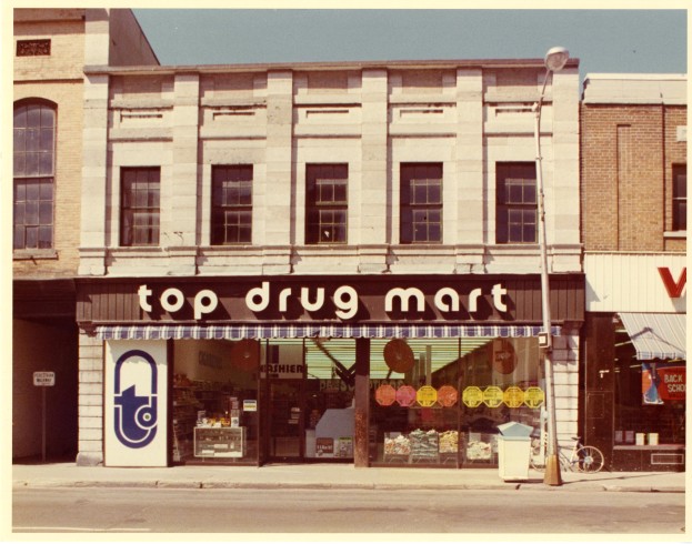 Eckladen namens "Top Drug Mart" mit einer Schautafel, Fenstern, einem Straßenschild, einem geparkten Fahrrad, einem Müllcontainer und einem bewölkten Himmel.