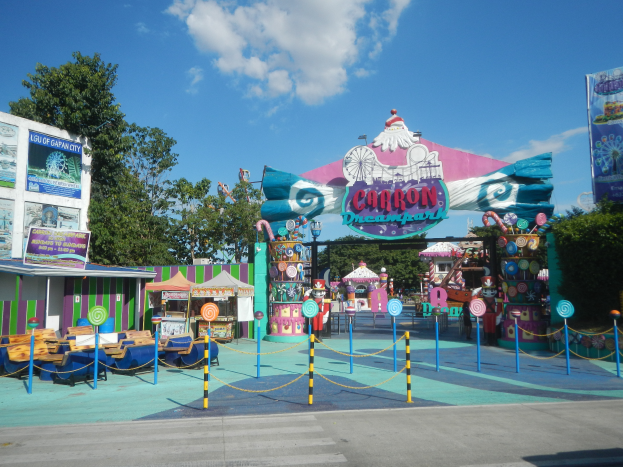 Eine Karussellfahrt am Eingang eines Parks mit einer Straße, einem Zaun, Buden, Schildern, Bäumen und einem bewölkten Himmel im Hintergrund.