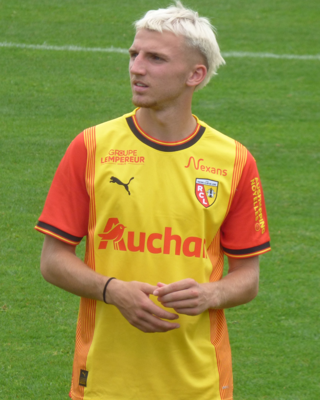 Ein Mann in einem gelben und orangefarbenen Fußballtrikot steht selbstbewusst auf einem Fußballfeld und sieht entschlossen aus.