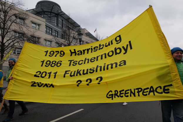 Gruppe von Menschen, die eine Straße mit einer gelben "Greenpeace"-Fahne entlanggehen, mit Bäumen und Himmel im Hintergrund.