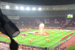 Ein Stadion mit Flutlicht, Eisengittern, Zuschauern, stehenden Personen, Werbetafeln, Bildschirmen, Überwachungskameras, Fahnen und Dekorationen.
