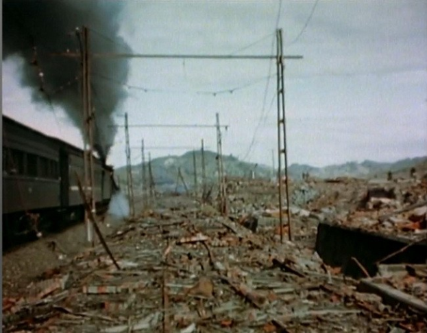 Ein Zug fährt durch ein beschädigt Gebiet mit Rauch, der aus ihm aufsteigt, umgeben von Strommästen und -drähten, mit Höhen und einem klaren Himmel im Hintergrund und verstreuten Trömmern am Boden.