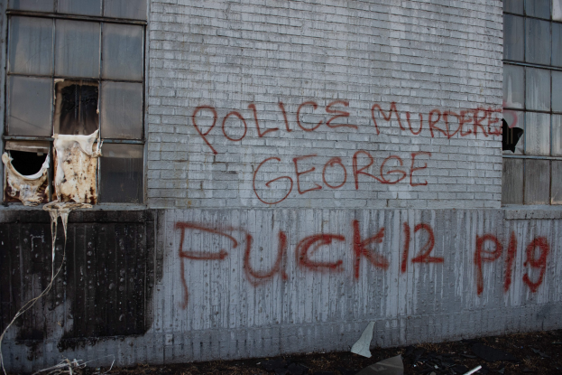 Ein Gebäude mit Graffiti, das "Polizei ermordet George Fuck 12 P19" auf der Seite trägt, mit Fenstern und diversen Gegenständen vorne.