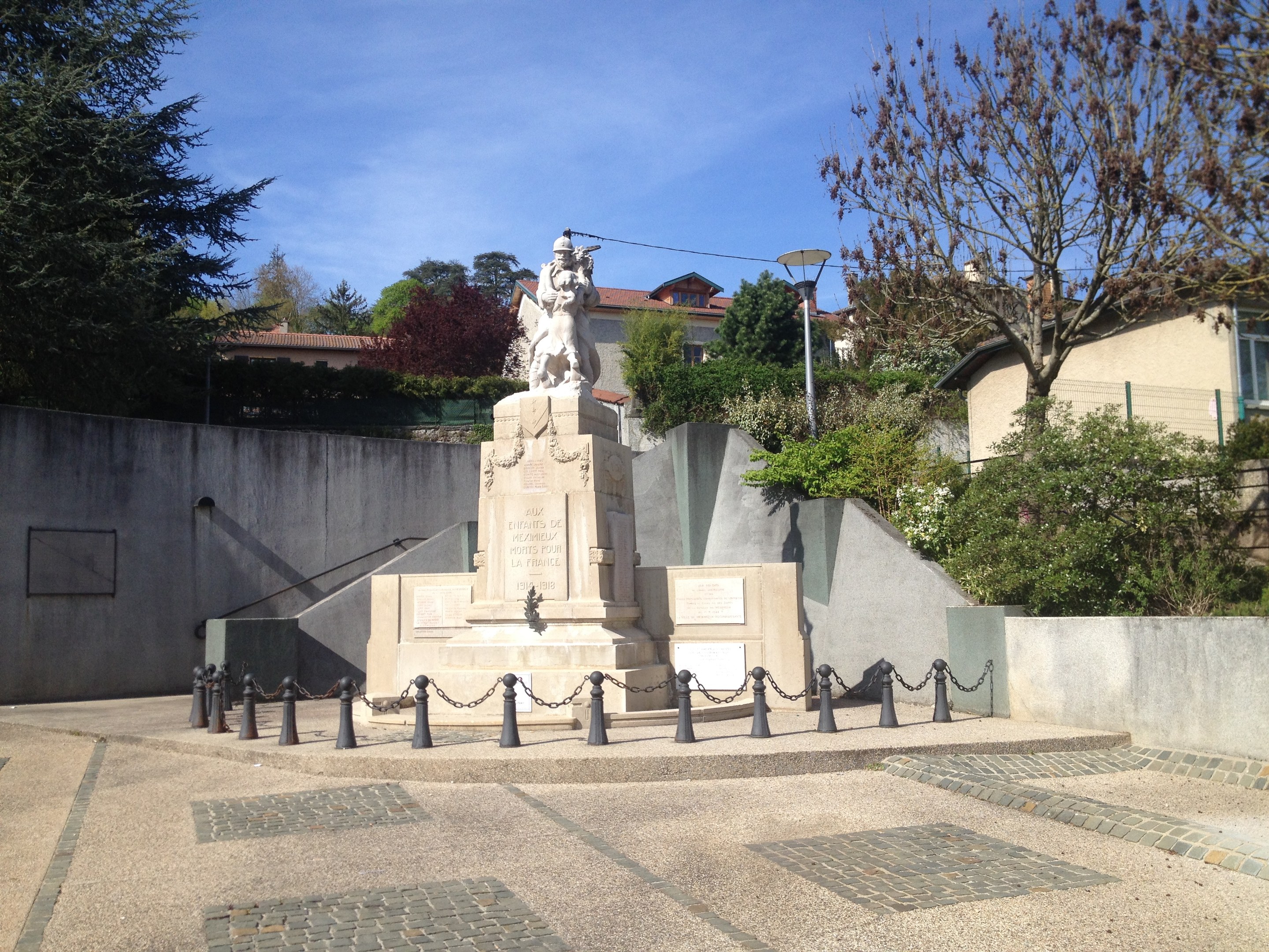 Eine Statue eines Mannes auf einem Sockel in einem Park, umgeben von einem Metallzaun, Bäumen, Pflanzen, einem Straßenmast, Gebäuden und einem bewölkten Himmel, die angeblich ein Denkmal für die Pariser Anschläge ist.