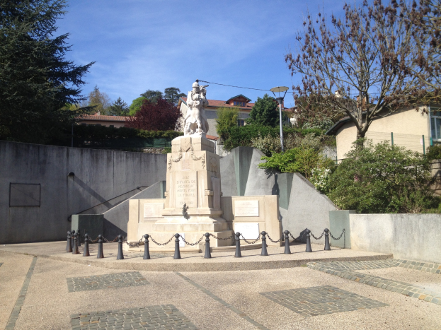 Eine Statue eines Mannes auf einem Sockel in einem Park, umgeben von einem Metallzaun, Bäumen, Pflanzen, einem Straßenmast, Gebäuden und einem bewölkten Himmel, die angeblich ein Denkmal für die Pariser Anschläge ist.