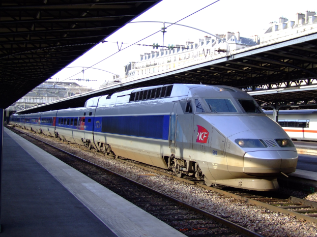 Hochgeschwindigkeitszug fährt auf Schienen neben einem Bahnsteig mit Gebäuden und einem klaren blauen Himmel im Hintergrund.
