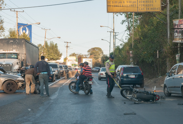 Gruppe von Menschen um ein verunglücktes Motorrad auf dem Straßenrand versammelt mit mehreren Fahrzeugen, einschließlich einem Lastwagen, im Hintergrund und Bäumen, Pfosten, Laternen, Schildern und Himmel sichtbar.