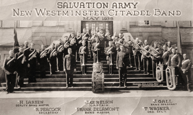 Gruppe von Menschen auf Treppenstufen stehend, die musikalische Instrumente halten, mit Fahnen und Pflanzen im Hintergrund, Text oben und unten lautet "Salvation Army New Westminster Citadel Band"