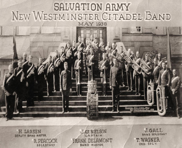 Gruppe von Menschen auf Treppenstufen stehend, die musikalische Instrumente halten, mit Fahnen und Pflanzen im Hintergrund, Text oben und unten lautet "Salvation Army New Westminster Citadel Band"