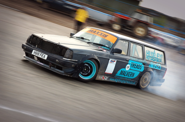 Ein Auto, das auf einer Rennstrecke fährt und Rauch hinter sich herzieht, umgeben von anderen Fahrzeugen und Zuschauern, mit einem unscharfen Hintergrund, der hohe Geschwindigkeit andeutet.