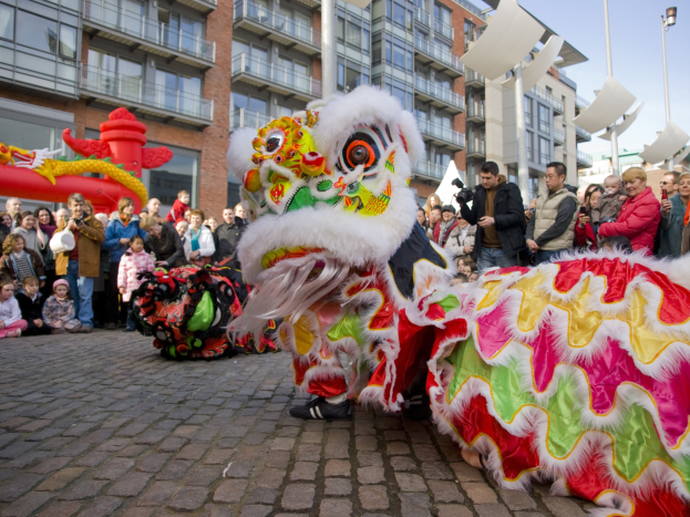 Ein lebendiges chinesisches Neujahrsfest in Amsterdam mit einer Drachenvorführung vor einer Zuschauermenge, einige fotografieren das Ereignis, vor einer Rückwand aus Gebäuden, Laternenmasten und einem klaren blauen Himmel.