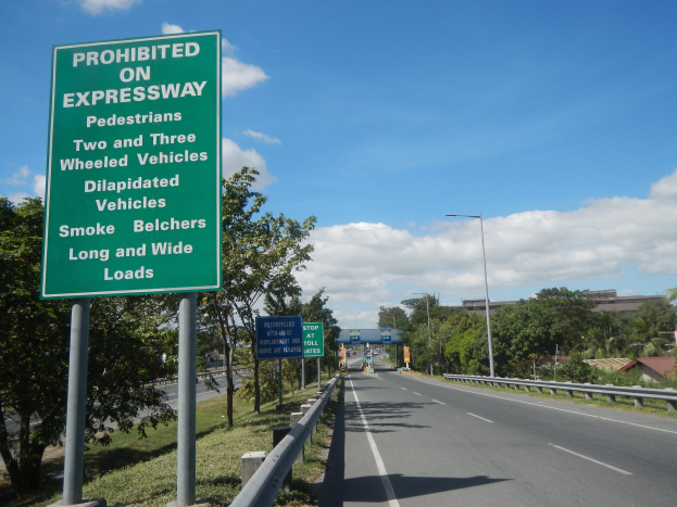 Grüne Highway-Schilder mit der Aufschrift "Verboten auf der Autobahn" mit Fahrzeugen, Laternenpfählen, Bäumen, Gebäuden und bewölktem Himmel im Hintergrund.