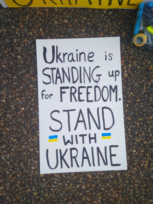 Ein weißer Papierbogen mit den Worten "Ukraine steht für Freiheit auf, stellt Euch mit Ukraine" liegt vor einem gelben Banner auf dem Boden. Auf der rechten Seite befindet sich eine Plastikhülle mit verschiedenen Gegenständen.