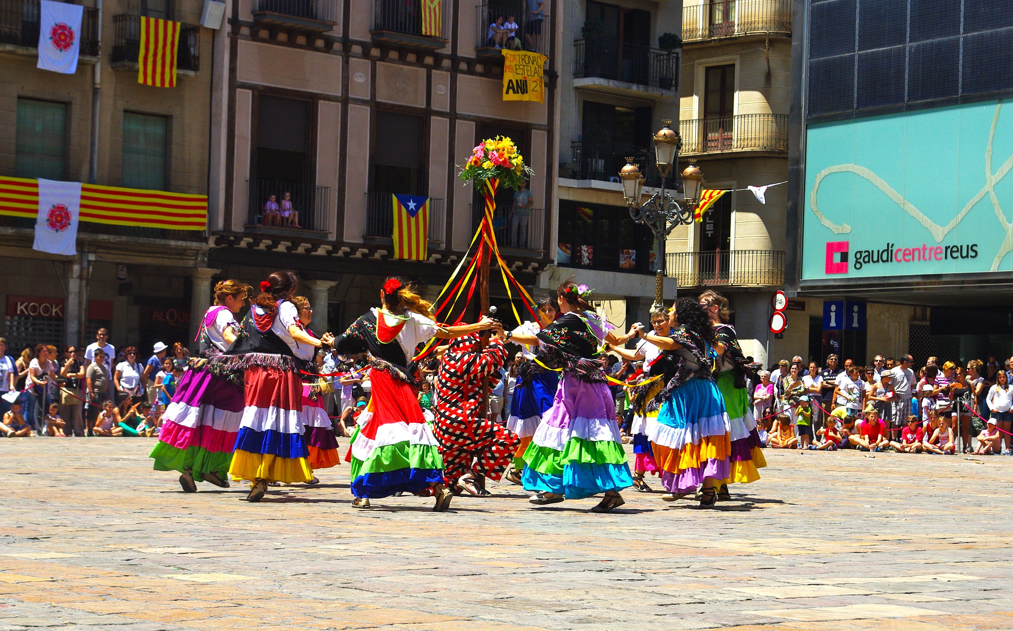 Eine Gruppe von Frauen in bunten Kleidern tanzt auf der Straße, umgeben von einer Menge Menschen, mit Gebäuden, Geländern, Balkonen, Fahnen, einem Lichtmast und einer Tafel mit Schrift im Hintergrund.