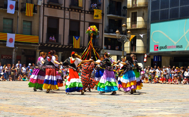 Eine Gruppe von Frauen in bunten Kleidern tanzt auf der Straße, umgeben von einer Menge Menschen, mit Gebäuden, Geländern, Balkonen, Fahnen, einem Lichtmast und einer Tafel mit Schrift im Hintergrund.