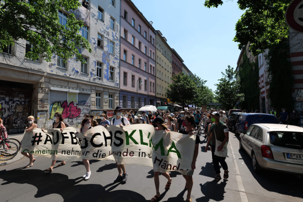 Eine Gruppe von Menschen marschiert auf einer Straße und hält ein Transparent, rechts geparkte Autos, links ein Fahrradfahrer, Gebäude, Bäume und ein Schild im Hintergrund, sichtbarer Himmel.