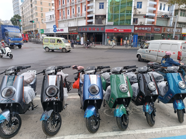 Eine Reihe von E-Scootern, die entlang einer Straße geparkt sind, mit einer Person in einer Mütze davor, Gebäuden, Verkehrsampeln, Bäumen und einem klaren Himmel im Hintergrund.