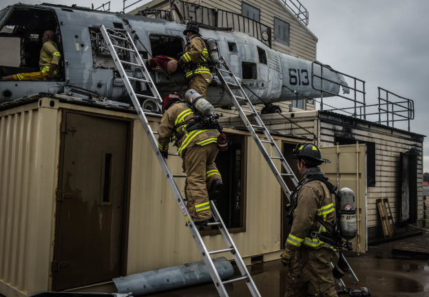 Feuerwehrleute in Uniform und Helmen arbeiten an einem Hubschrauber auf einem Gebäudedach, mit Leitern, einem Sauerstoffzylinder, einem nahen Container, Bäumen und einem klaren blauen Himmel im Hintergrund.