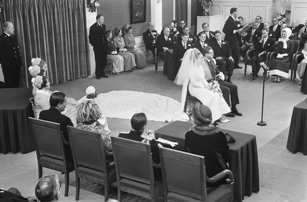 Schwarzes und weißes Foto einer Hochzeitszeremonie mit sitzenden und stehenden Menschen, einem Mann, der in ein Mikrofon spricht, und einer Frau, die einen Strauß hält, mit einem Rednerpult, Blumenschmuck, einem Vorhang, einem gerahmten Foto an der Wand und einer Tür im Hintergrund.
