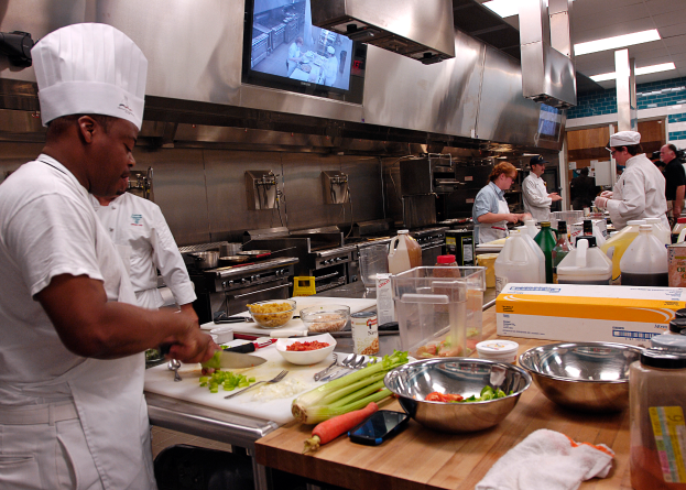 Eine Gruppe von Köchen in einer Küche bei der Zubereitung von Speisen, mit einem Koch im Vordergrund in einer weißen Schürze und Mütze, der ein Messer hält, und umgeben von verschiedenen Utensilien, Schüsseln, Gemüse, einem Mobiltelefon, Flaschen und anderen Gegenständen, mit einem Fernseher und Deckenleuchten im Hintergrund.