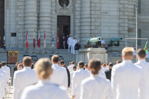 Gruppe von Menschen in weißen Marineuniformen vor einem Gebäude mit Säulen, Fahnen, einem Podium und Kanonen im Hintergrund.