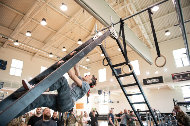 Ein Mann führt Klimmzüge an einer Leiter in einem Fitnessstudio, während ihn eine Gruppe von Menschen mit Kameras beobachtet; Fitnessgeräte und Banner sind im Hintergrund zu sehen.