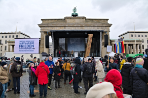Eine Menschenmenge steht vor einem Gebäude mit einer Bühne, auf der Lautsprecher und ein Bildschirm zu sehen sind, sowie Fahnen und Schilder auf der rechten Seite, was auf eine Demonstration in Berlin hindeutet.