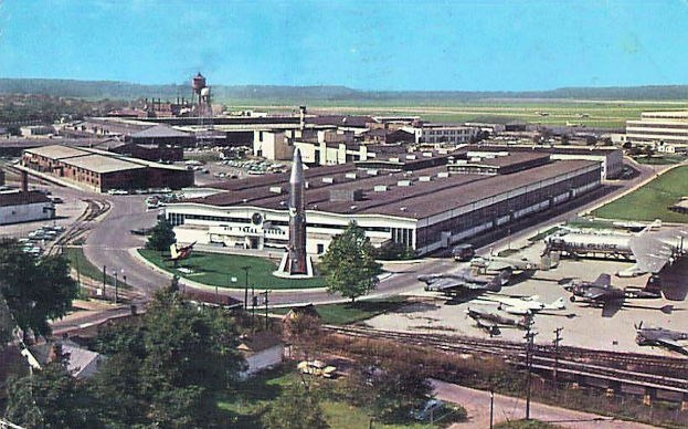 Altes Schwarz-Weiß-Foto eines Flughafens mit mehreren Propellerflugzeugen auf dem Rollfeld geparkt, umgeben von Gebäuden, Bäumen, Straßen, Fahrzeugen, Strommasten, Grasflächen, Bahnschienen, Türmen, fernen Hügeln und einem klaren blauen Himmel, mit Text unten.