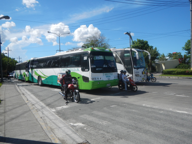 Ein grüner und weißer Shuttlebus steht am Straßenrand mit Motorradfahrern davor, ein grasbewachsener Fußweg links daneben und Gebäude, Bäume, Laternenpfähle und einen klaren blauen Himmel im Hintergrund.