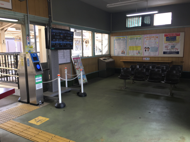Ein belebter Bahnhofseingang mit Sitzgelegenheiten, einer Informations counter, Ticketautomaten, Deckenleuchten, Geländern und Informationsanzeigen.