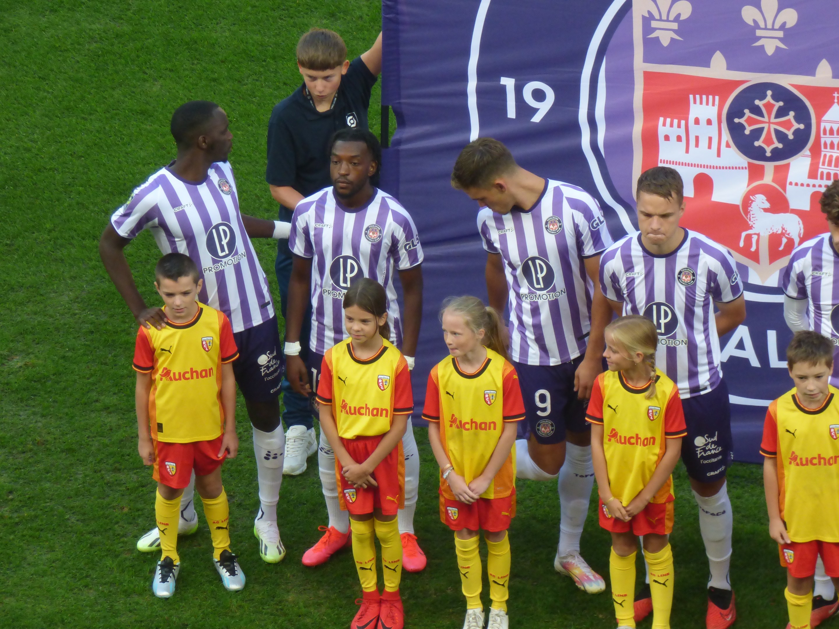 Eine Gruppe junger Menschen in Sportkleidung steht auf einem saftig grünen Fußballfeld mit einem "Ligue 1"-Plakat im Hintergrund.