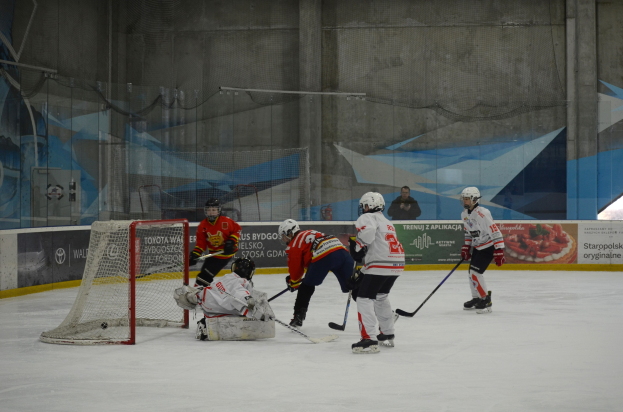 Eine Gruppe von Menschen, die Eis hockey auf einem Eisplatz spielt, trägt Helme und hält Hockey-Schläger, mit einem Torpfosten auf der linken Seite und Bannern im Hintergrund.