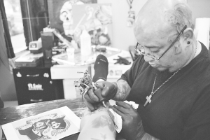 Ein Schwarz-Weiß-Bild eines Mannes in einem schwarzen T-Shirt, mit Brille und einer Kette um den Hals, der mit einer Maschine ein Bein tätowiert, mit einem Papier auf der Oberfläche und einem unscharfen Hintergrund mit einer Flasche, einem Becher und anderen Gegenständen.