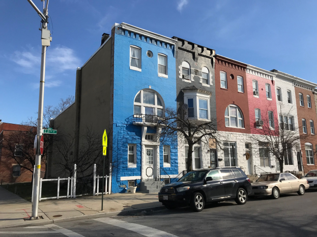 Eine Reihe bunter Häuser säumt eine Straßenkreuzung mit parkenden Autos, Bäumen, Strommasten, einem Zaun und einem bewölkten Himmel.