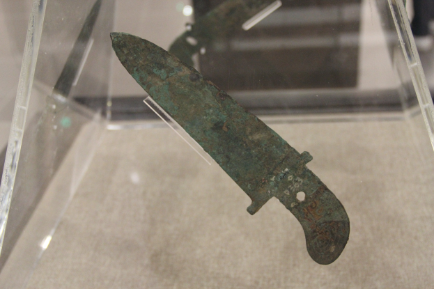 Ein Messer in einer durchsichtigen Glasvitrine mit detaillierter Klingenansicht, vor einem unscharfen Museums-Hintergrund.