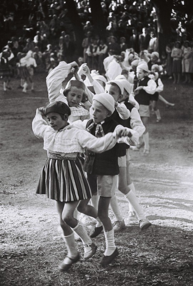 Schwarzes und weiße Foto von Kindern, die in einem Park tanzen, mit einigen Zuschauern und Bäumen im Hintergrund.