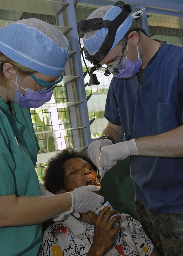 Zwei Chirurgen in Masken und Handschuhen führen eine Gesichtsoperation an einer Frau durch, die auf einem Bett liegt, während durch ein Fenster im Hintergrund Bäume zu sehen sind.