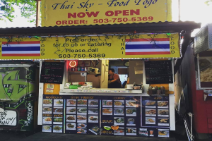 Eine Geschäftsfront mit verschiedenen Gegenständen, darunter Getränkedosen, Speisekarten, eine Uhr, Gewürze in Plastikbehältern, Gewürze und Werbetafeln unter einem klaren Himmel.