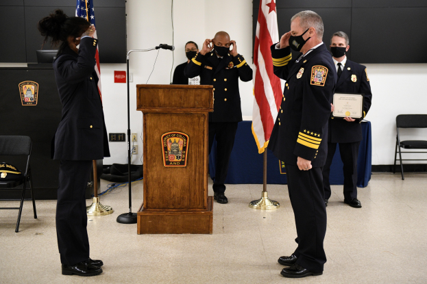 Eine Gruppe uniformierter Personen in Masken steht vor einem Podium mit einem Mikrofon, umgeben von Flaggen, Stühlen, Tischen und einer Wand, wobei eine Person ein Zertifikat hält, während es apparently eine Vereidungszeremonie für einen neuen Feuerwehrchef gibt.