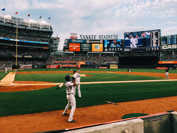 Ein Baseball-Spiel an einem bewölktem Tag im Yankee Stadium mit Spielern auf dem Feld und Zuschauern in den Rängen, umgeben von Stadionmerkmalen wie Zäunen, Fahnen, Anzeigetafeln, einem Display und Deckenleuchten.
