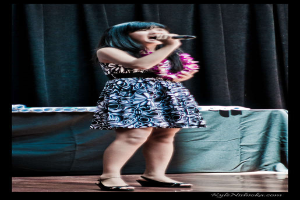 Eine Frau in einem bunten Kleid singt in ein Mikrofon auf der B├╝hne, mit einem Tisch mit Papieren und einem Vorhang im Hintergrund.